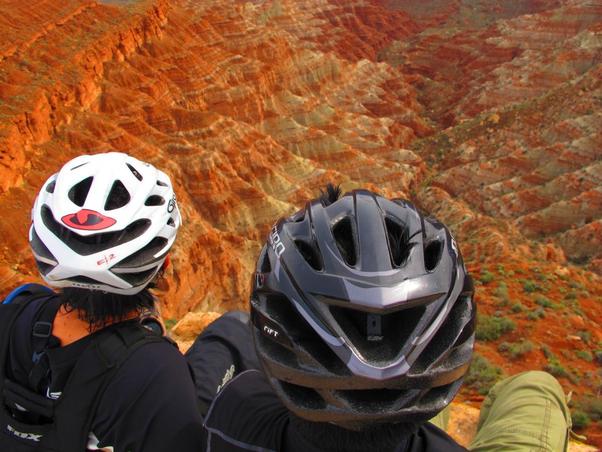 Two cyclists resting at the edge of a colorful canyon, wearing white and black helmets, with a stunning view of layers of red and orange rock formations in the background. Gooseberry Mesa mountain bike trail.