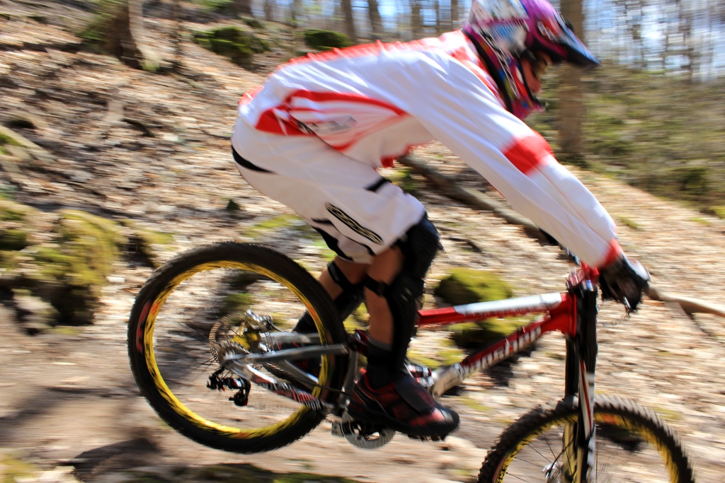 A mountain biker in motion, wearing protective gear and a colorful helmet, jumps over a rocky section of a trail surrounded by trees and foliage. The image captures the speed and excitement of the sport with motion blur emphasizing the bike's movement.
