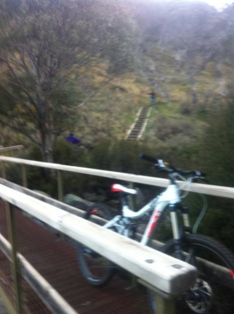 A mountain bike leaning against a wooden railing, with a gravel path and a set of stairs visible in the background, surrounded by trees and greenery. A person in a purple jacket can be seen walking down the path. Dead Horse Gap mountain bike trail.