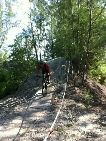 A mountain biker in a red shirt rides down a dirt trail on a slope surrounded by trees. The path is lined with a white rope, and the sun filters through the foliage, creating a natural, outdoor setting. Amelia Earhart Park mountain bike trail.