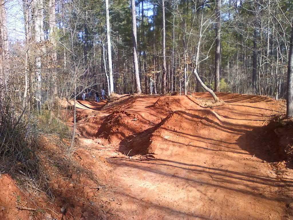 A scenic view of a dirt bike track winding through a forested area. The path features mounds of reddish dirt and curves, with tall trees lining either side. Some people can be seen in the background, indicating activity in the area. The ground is bare, showing signs of recent excavation or construction. Quehl Holler mountain bike trail.
