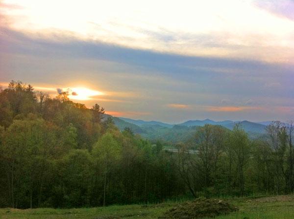 A scenic landscape view at sunset, featuring rolling hills and a forested foreground. The sky is painted with soft hues of orange and pink as the sun sets behind the mountains, creating a tranquil atmosphere. Rocky Knob Park mountain bike trail.