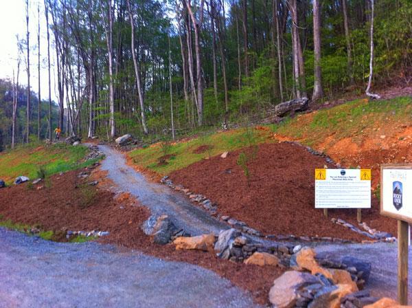 A gravel pathway leads into a forested area, flanked by newly cleared land and lined with rocks. On the right, a sign warns of trail conditions, and trees with fresh green foliage can be seen in the background, indicating the onset of spring or early summer. Rocky Knob Park mountain bike trail.
