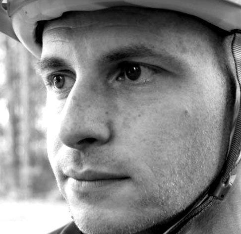 A close-up, black-and-white portrait of a man wearing a helmet, gazing thoughtfully into the distance. His expression is focused, highlighting his features such as freckles and the contours of his face. A blurred natural background is slightly visible behind him.