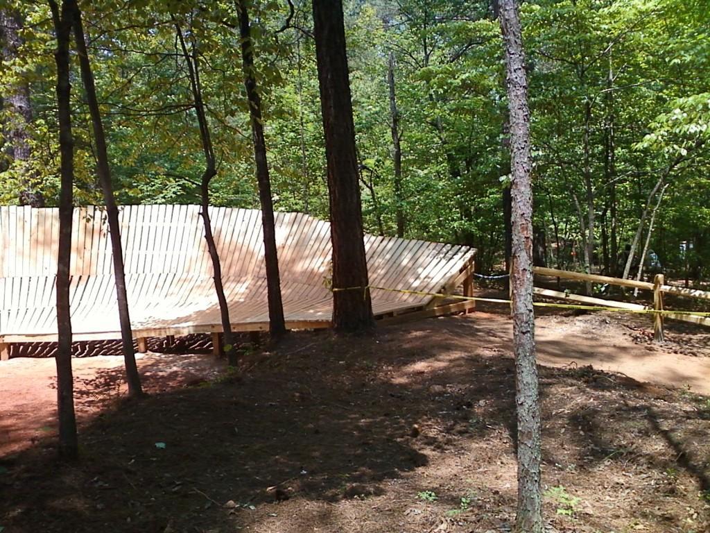 A wooden structure resembling a ramp, located in a forested area with green trees and a dirt path. The ramp is partially constructed and secured with yellow caution tape. Quehl Holler mountain bike trail.