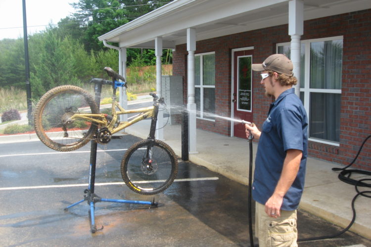 A man in a blue shirt and shorts is washing a muddy yellow mountain bike, which is mounted on a repair stand. He is using a hose to spray water on the bike, which is situated outside a brick building in a parking area. Green trees are visible in the background, and the ground is wet from the water.