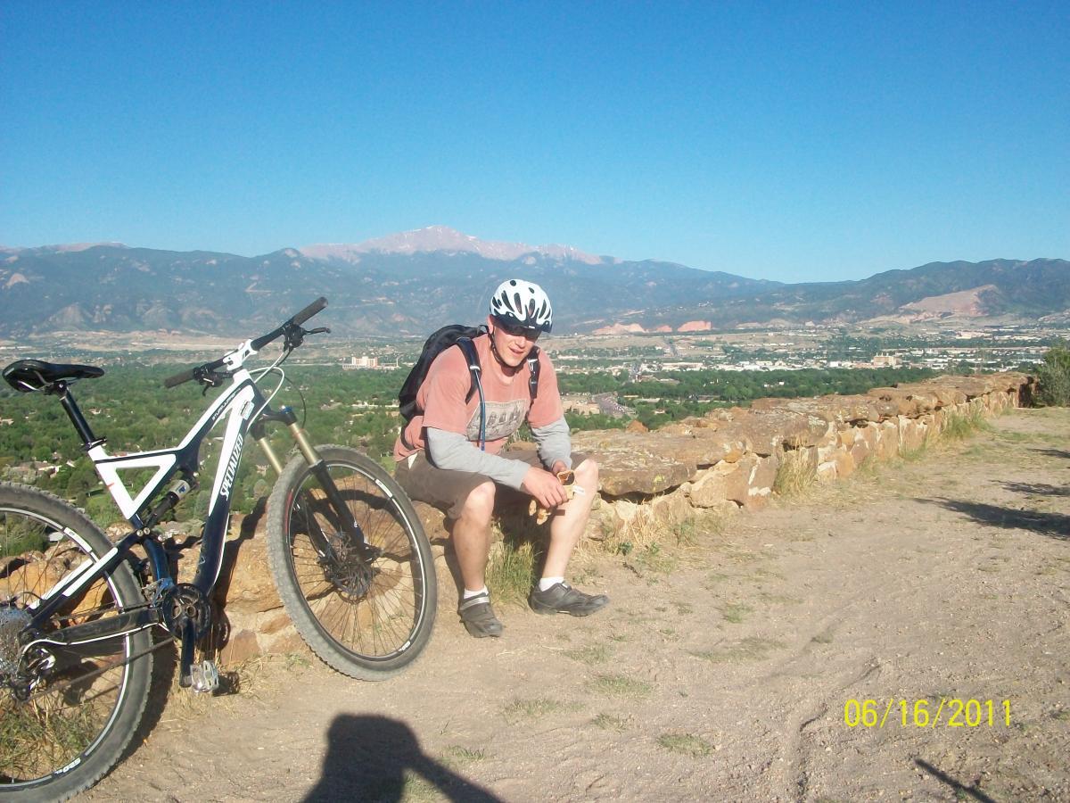 A person sitting on a rocky ledge, resting during a mountain biking trip, with a mountain bike positioned nearby. The background features a clear blue sky and a panoramic view of hills and valleys. The individual is wearing casual clothing, a helmet, and appears to be enjoying the scenery. Palmer Park mountain bike trail.