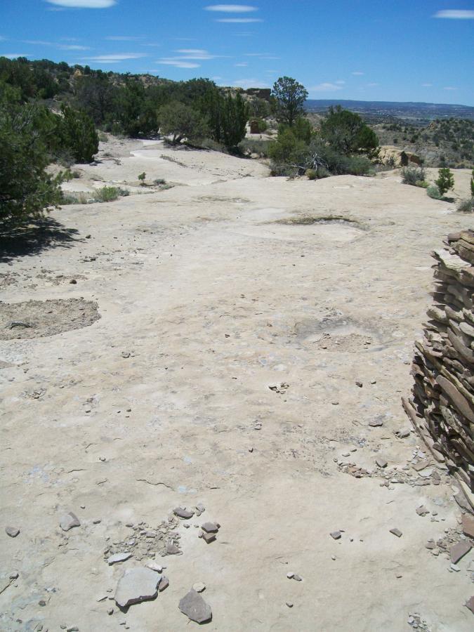 A dry, rocky landscape under a blue sky with scattered clouds. The foreground features a sandy terrain with small stones and patches of vegetation. In the background, there are low shrubs and distant hills. A stone wall is visible on the right side of the image, suggesting a historical or archaeological site. High Desert Trail System mountain bike trail.