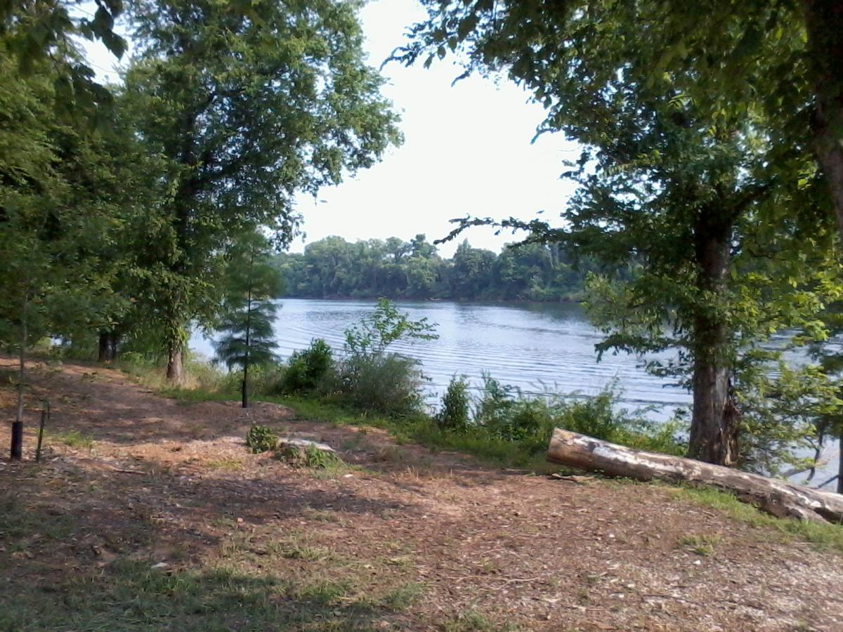 A scenic view of a riverbank surrounded by lush greenery. Trees frame the image, with a calm river in the background reflecting the clear sky. The foreground features a natural path with scattered stones and patches of grass, creating a serene, outdoor atmosphere. North Augusta Greeneway mountain bike trail.