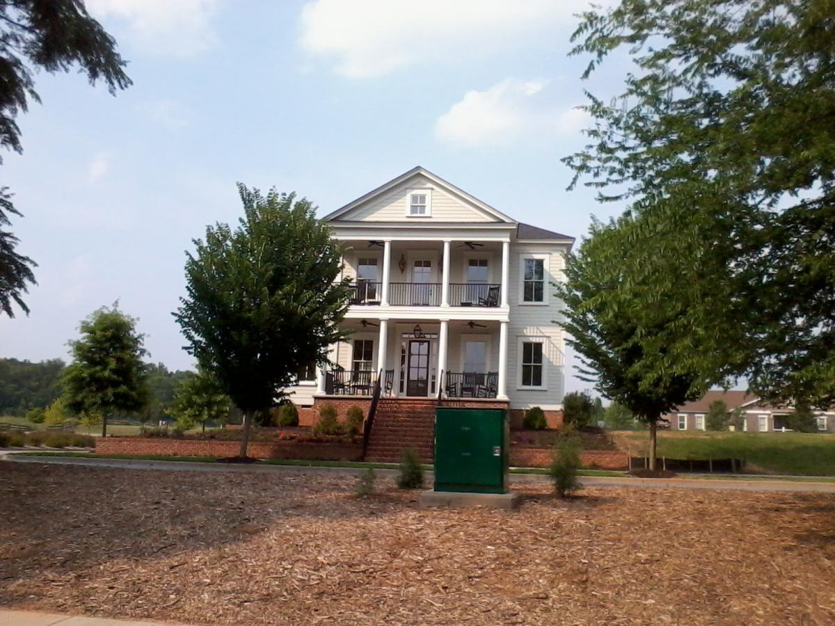 A two-story white house with a gabled roof, featuring a spacious front porch with rocking chairs, surrounded by green trees and landscaping. The house is set against a blue sky with a few clouds, and there is a green utility box near the front. North Augusta Greeneway mountain bike trail.