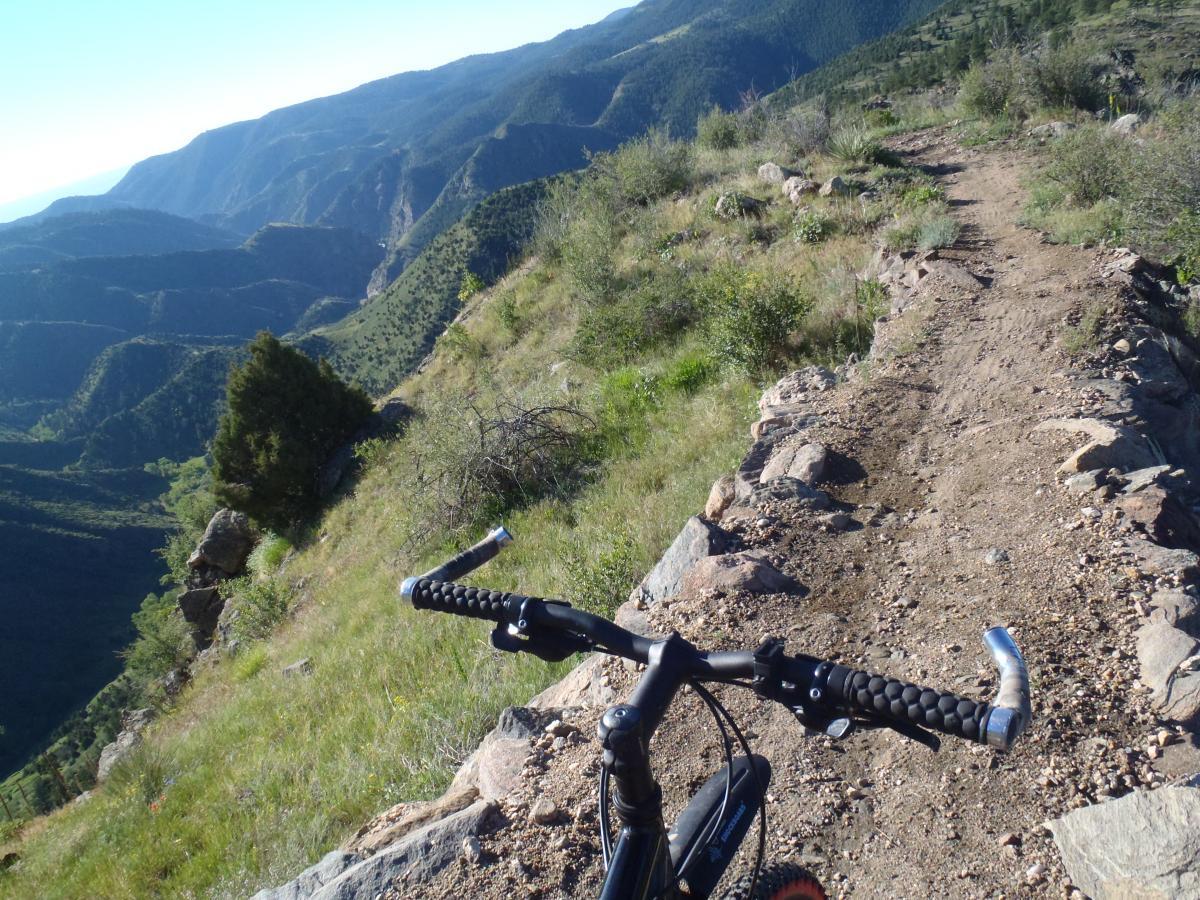 A mountain bike on a rocky trail overlooking a lush green valley and distant mountains under a clear blue sky. The handlebars of the bike are visible in the foreground, with rugged terrain on both sides of the path. Centennial Cone Park mountain bike trail.