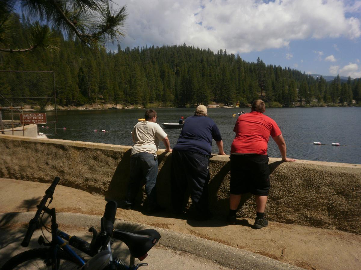 Three people are leaning against a stone wall by a lake, looking out at the water. The scene is surrounded by lush green trees and a partly cloudy sky. A bicycle is parked on the ground nearby. A sign warns of a dangerous area beyond the wall. Hume Lake Trail mountain bike trail.