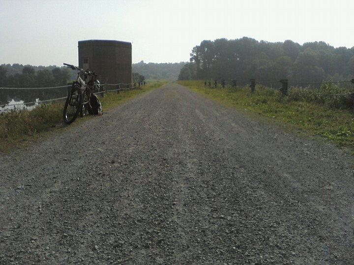 A gravel path beside a body of water, with a bicycle parked on the left and a small building in the background. Lush greenery and trees line the path, creating a serene, natural setting on a clear day. Marsh Creek Park mountain bike trail.