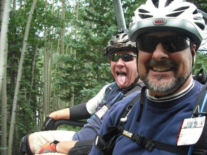 Two men wearing helmets and sunglasses are sitting on a ski lift, surrounded by greenery. One man is smiling broadly, while the other is playfully sticking out his tongue. Both are dressed in casual athletic wear and appear to be enjoying their outdoor adventure. Sunrise mountain bike trail.