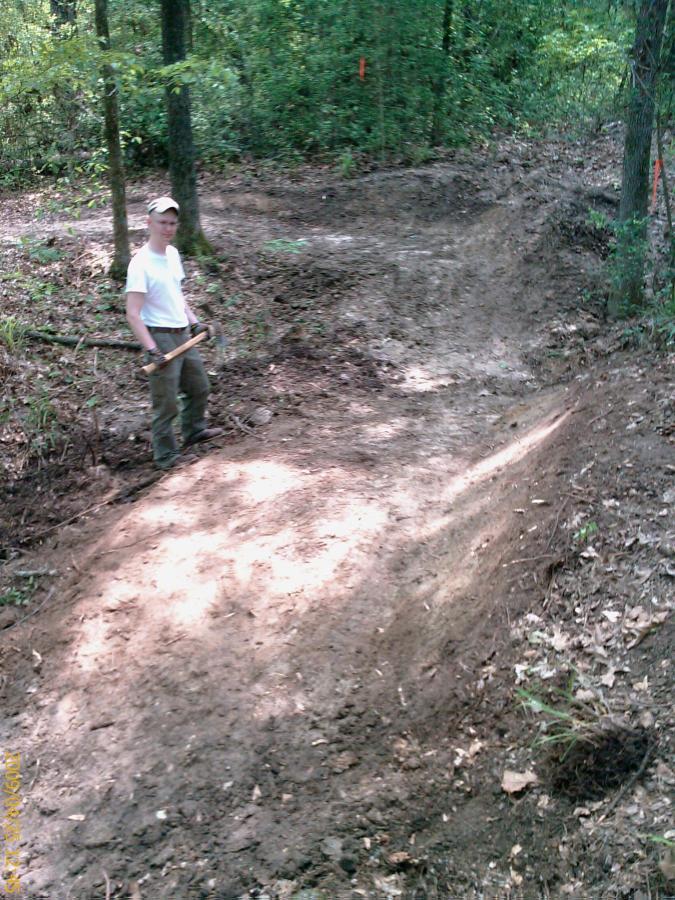 A person standing on a dirt trail in a wooded area, holding a tool. The trail shows signs of recent digging and construction, surrounded by trees and greenery. Kit Mcconnico Lost Arrow mountain bike trail.