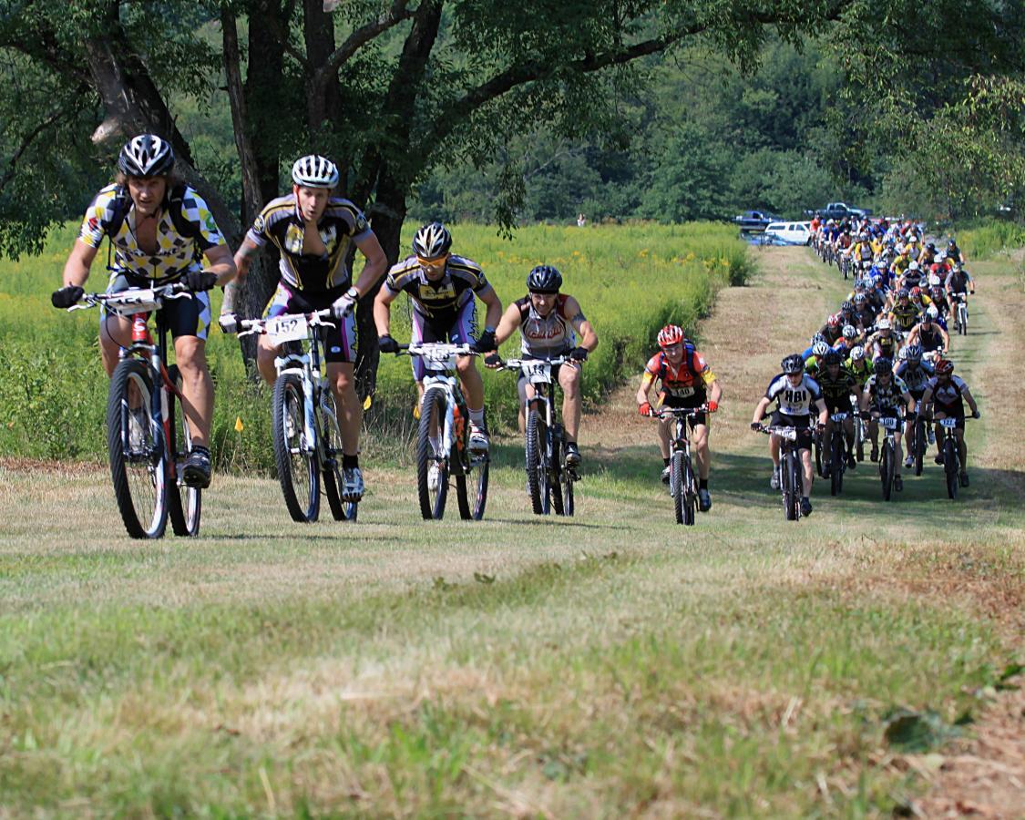 A group of mountain bikers racing along a grassy trail surrounded by trees and wildflowers. The scene captures several riders in colorful jerseys and helmets, with the nearest cyclists appearing focused and determined as they navigate the course. A long line of additional riders can be seen in the background, indicating a larger cycling event. Two Mile Run County Park mountain bike trail.
