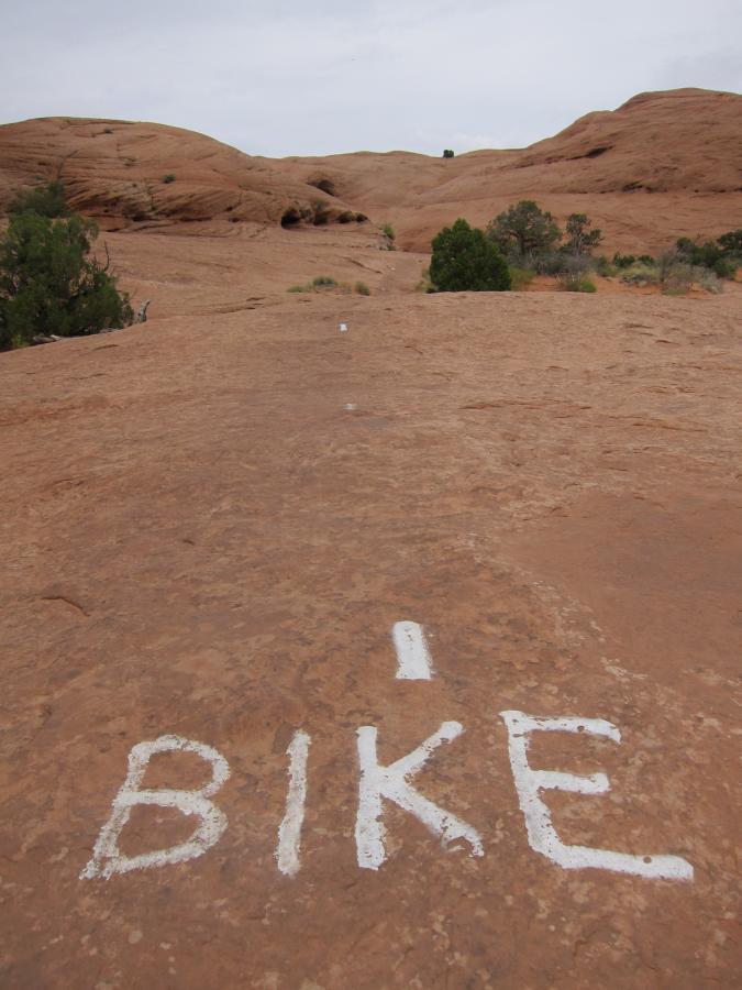 A rocky landscape with reddish-brown terrain, featuring large, smooth rock formations and sparse vegetation. In the foreground, white paint spells out "I BIKE" on the ground, indicating a bikers' path. The sky is overcast, and the overall scene suggests a natural outdoor area suitable for biking. Slickrock mountain bike trail.