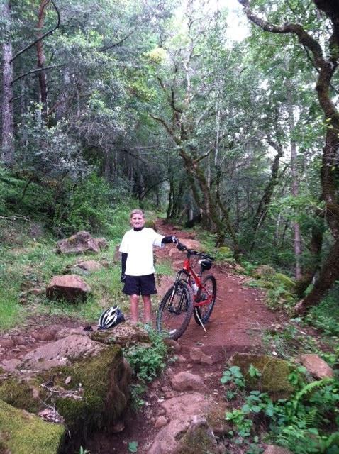 A young boy stands on a dirt path in a forested area, smiling and holding onto his bicycle. He is wearing a white t-shirt and shorts, with a helmet resting beside him. The surroundings are lush with greenery, including tall trees and scattered rocks along the trail. Annadel State Park mountain bike trail.