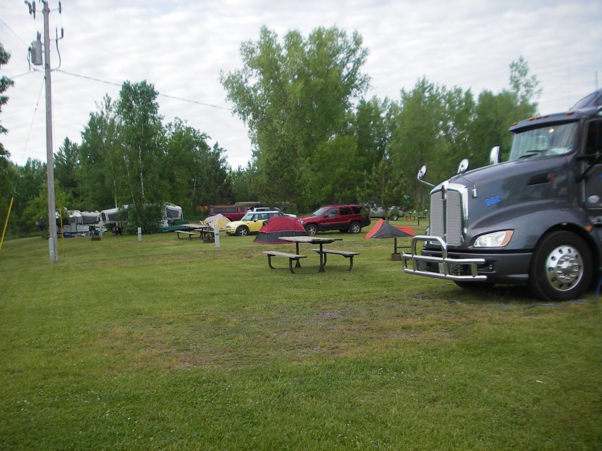 A grassy area in a campsite featuring a large gray truck parked in the foreground. Several picnic tables are arranged on the lawn, with two tents set up in the background. Various vehicles, including a red SUV and a yellow car, are parked nearby, surrounded by trees under a cloudy sky. Cuyuna Lakes mountain bike trail.