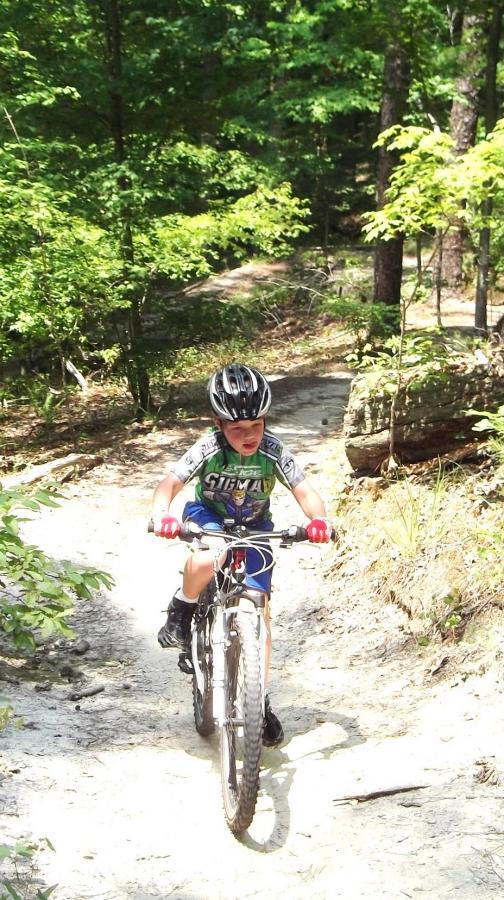 A young child wearing a helmet and bicycle gear rides a mountain bike along a dirt trail in a wooded area. Sunlight filters through the trees, highlighting the child focused on navigating the path amidst greenery. Harbison State Forest mountain bike trail.