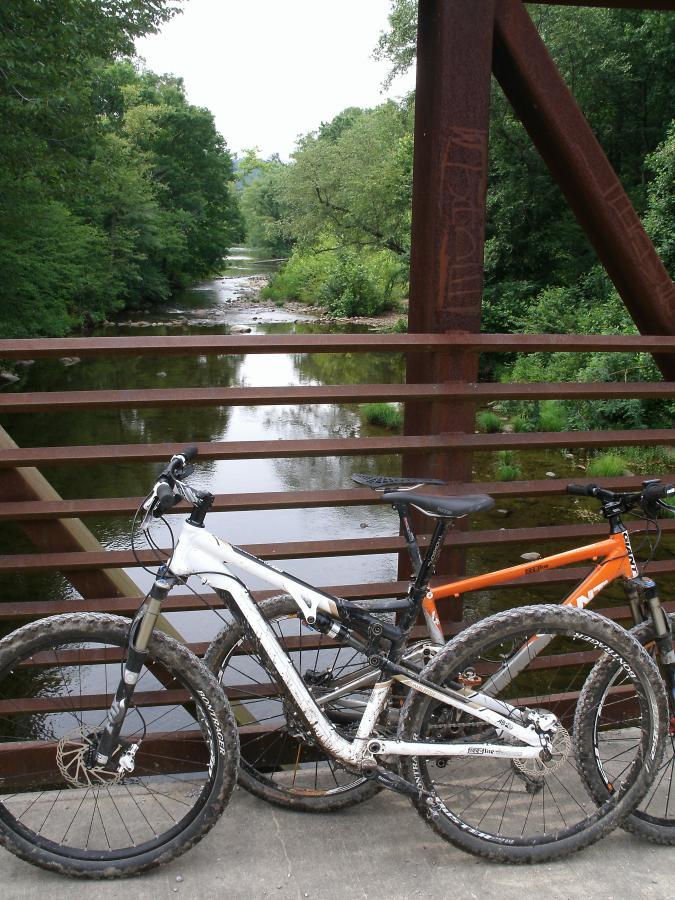 Two mountain bikes, one white and one orange, are resting against a railing on a metal bridge overlooking a serene stream surrounded by lush greenery. The scene captures a peaceful outdoor environment, ideal for biking and enjoying nature. Iron Mountain Loop mountain bike trail.