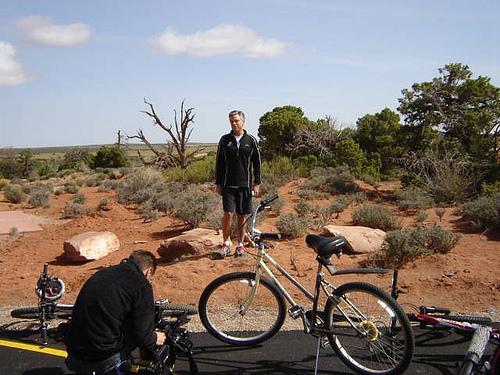 A man standing near a bicycle on a rugged landscape with red soil and sparse vegetation. Another individual is seen working on a bike in the foreground. The scene is set against a backdrop of trees and a partly cloudy sky. Dead Horse Point State Park mountain bike trail.