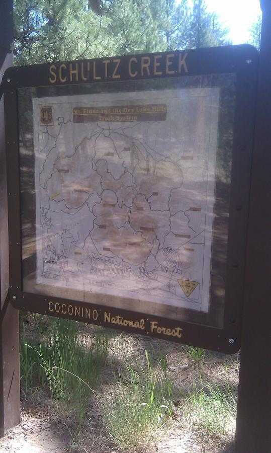 Sign for Schultz Creek Trail in Coconino National Forest displaying a map of the Mt. Elden and Dry Lake Hills Trail System, surrounded by green grass and trees. Schultz Creek mountain bike trail.