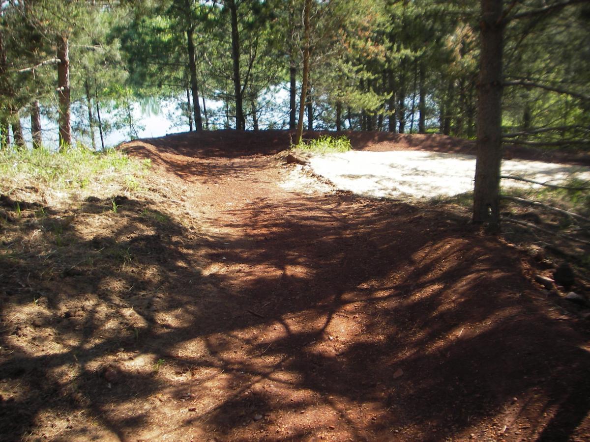 A winding dirt path surrounded by trees, leading towards a body of water in the background. Cuyuna Lakes mountain bike trail.