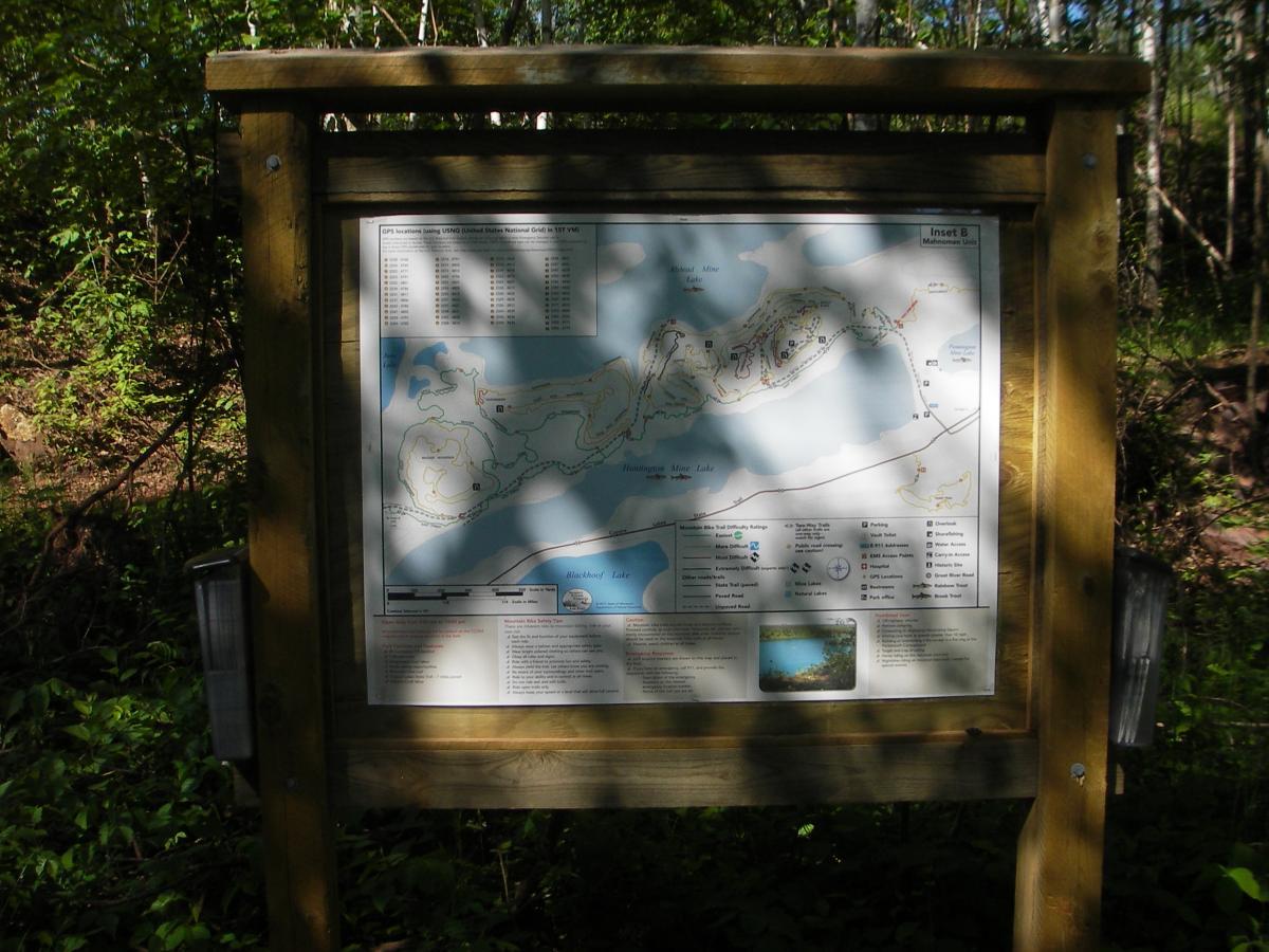 A wooden sign displaying a detailed map of a recreational area, surrounded by lush greenery. The map includes trails, water bodies, and various facilities, with a legend at the bottom for navigation. Sunlight filters through the trees, casting shadows on the map. Cuyuna Lakes mountain bike trail.