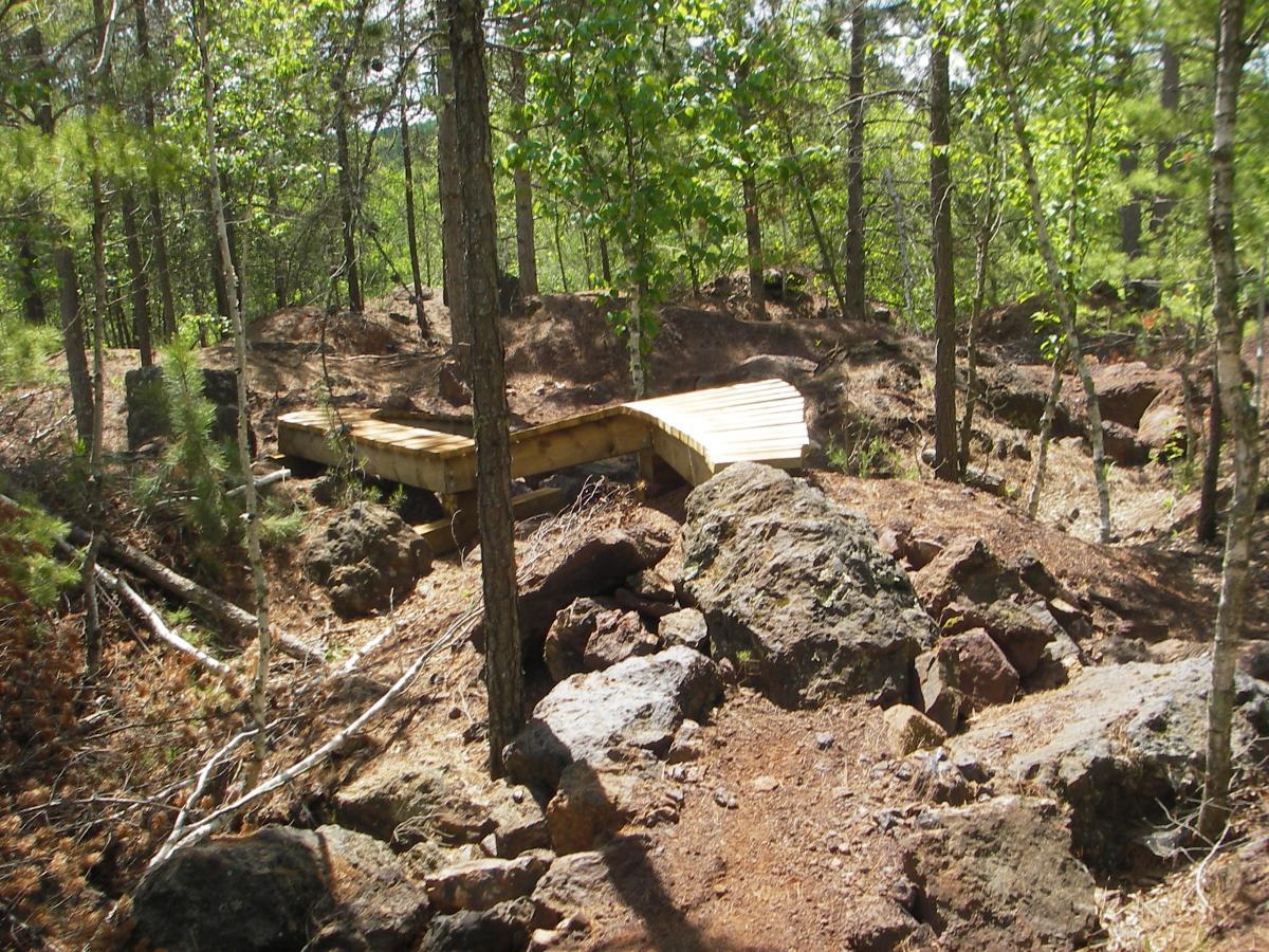 A wooden bridge spans a rocky area in a forested landscape, surrounded by tall trees and exposed roots. The path is earthy with scattered stones and vegetation, providing a natural setting for outdoor activities. Cuyuna Lakes mountain bike trail.