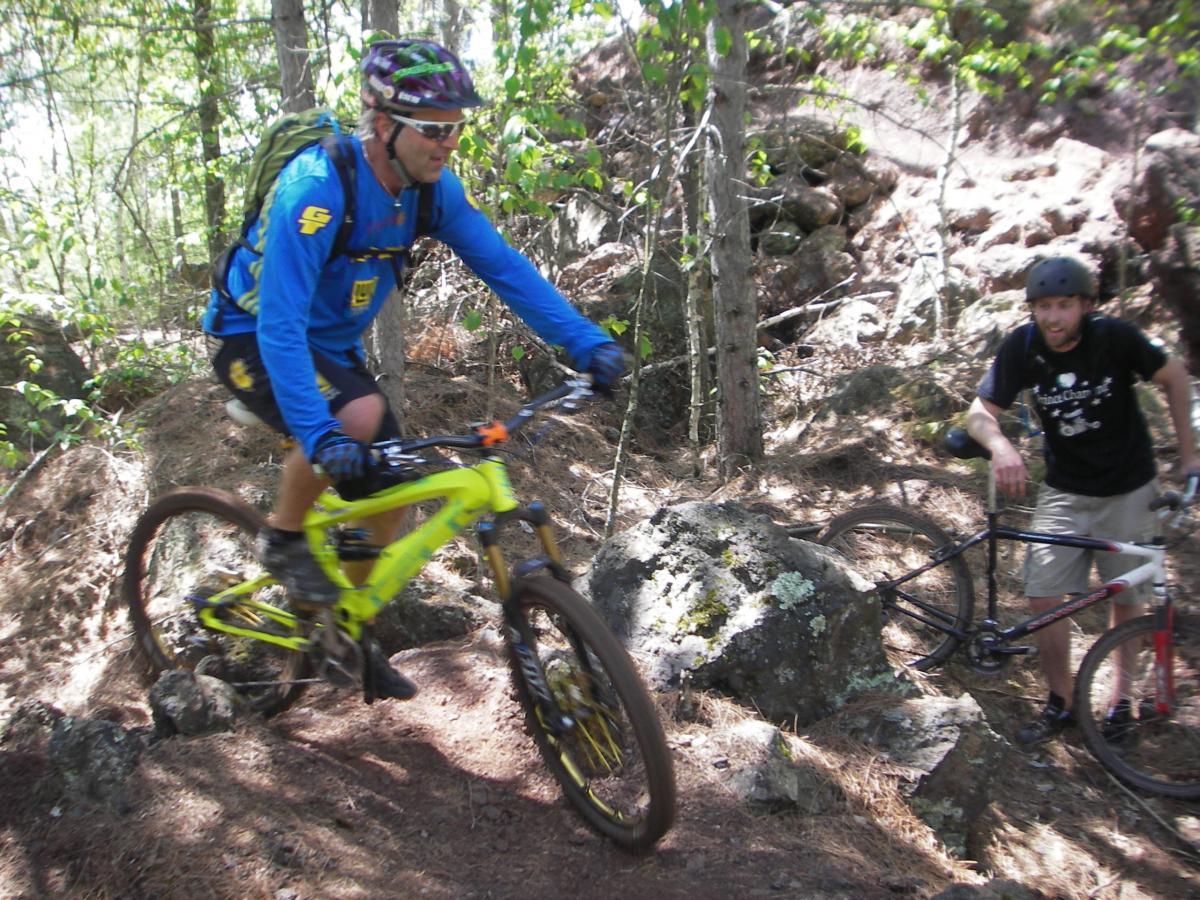 Two mountain bikers navigating through a rocky, forested trail. One rider, wearing a blue shirt and helmet, is actively cycling over a rocky section, while the other, in a black shirt and helmet, stands beside his bike, observing. Lush greenery surrounds them, providing a vibrant outdoor setting. Cuyuna Lakes mountain bike trail.
