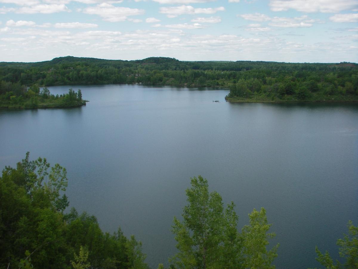 A serene view of a calm lake surrounded by lush greenery and rolling hills, under a partly cloudy blue sky. The still water reflects the landscape, creating a peaceful and tranquil atmosphere. Cuyuna Lakes mountain bike trail.
