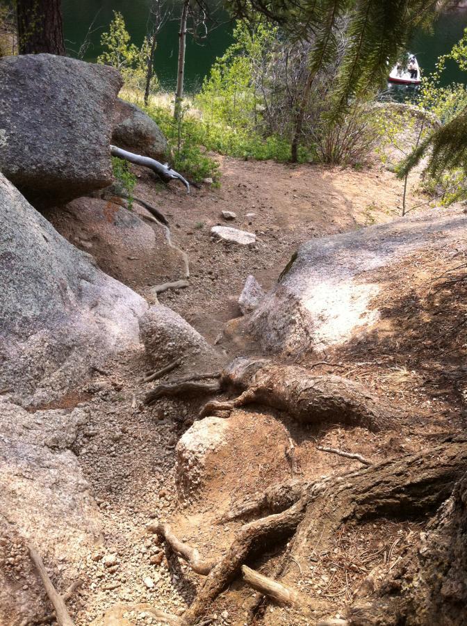 A dirt path winding between large rocks, surrounded by greenery and trees, leading down to a serene body of water in the background. A small boat is visible on the water. Rampart Reservoir mountain bike trail.