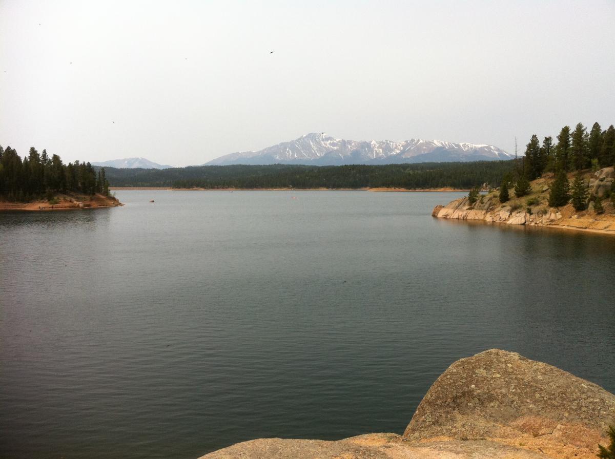 A serene landscape depicting a calm lake surrounded by forests and mountains. The water reflects the overcast sky, while snow-capped peaks in the background tower over the scene. A rocky shoreline is visible in the foreground, adding to the natural beauty of the setting. Rampart Reservoir mountain bike trail.