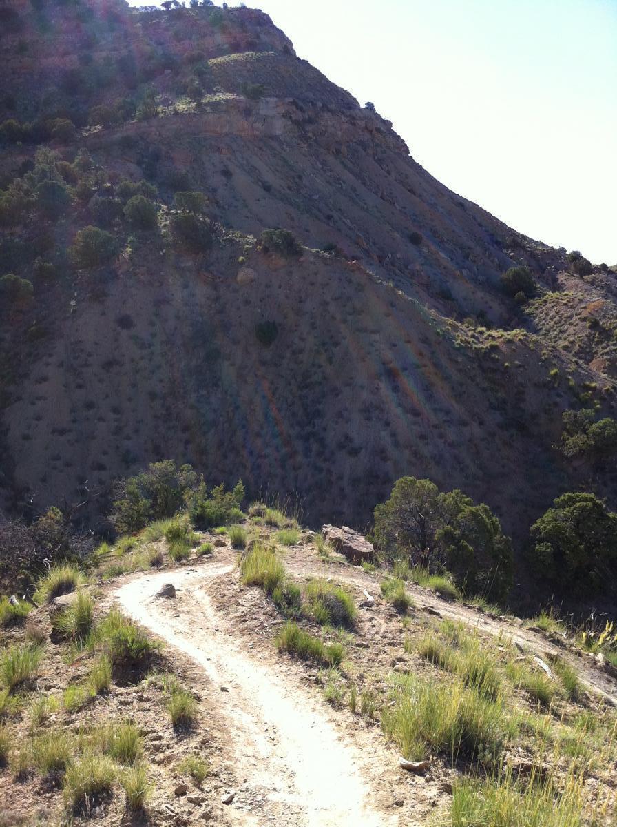 A winding dirt trail leads along a hillside with sparse vegetation and small shrubs. The background features steep, rocky slopes under a clear blue sky, suggesting a natural outdoor setting ideal for hiking or exploring. Chutes and Ladders mountain bike trail.