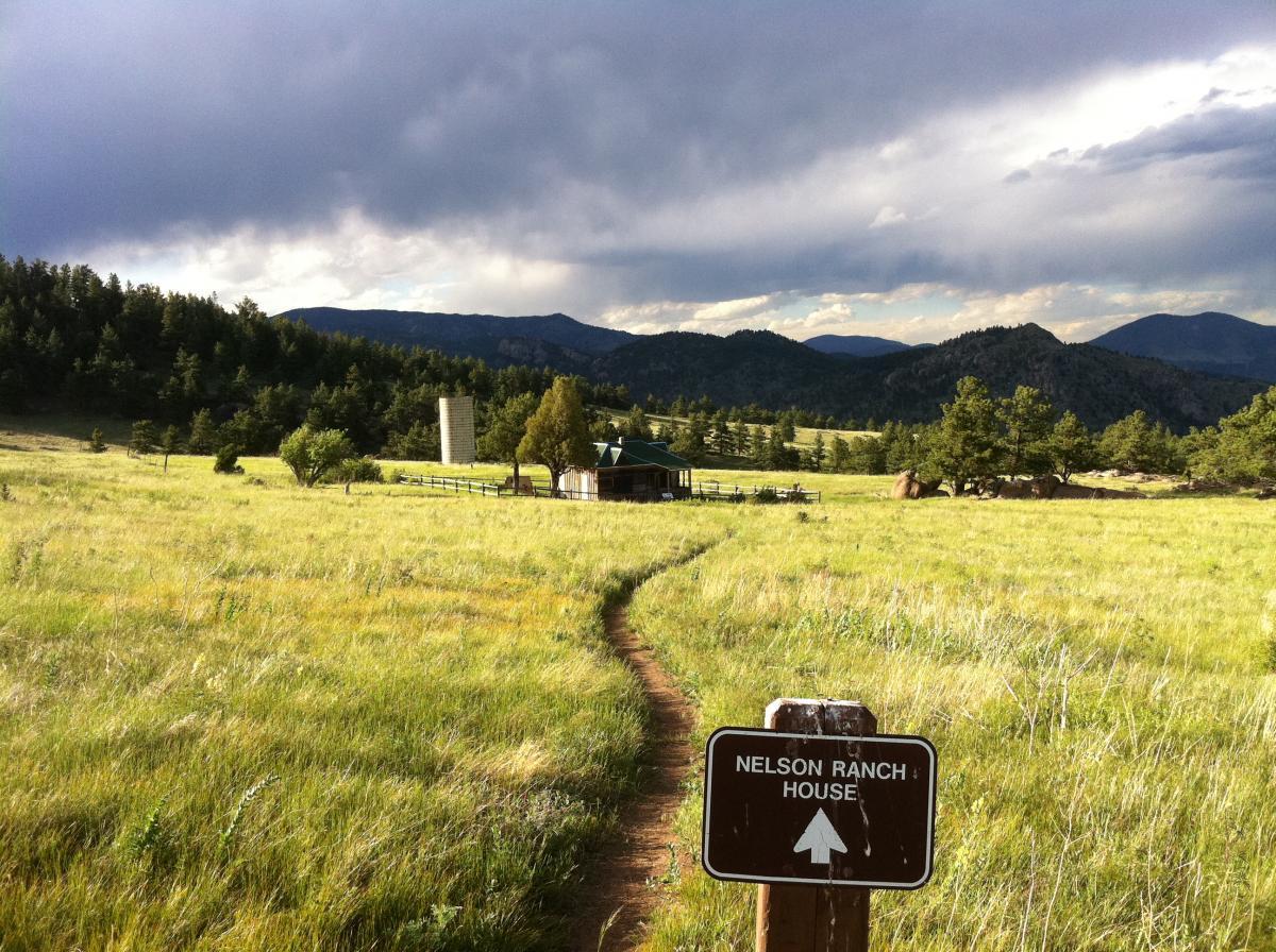 A dirt path leads through a grassy field toward a rustic wooden ranch house surrounded by trees, with a silo visible nearby. In the foreground, a sign indicates "Nelson Ranch House" with an arrow pointing forward. The background features rolling hills and a cloudy sky. Hall Ranch mountain bike trail.