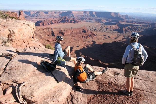 Three cyclists relax on a rocky outcrop, overlooking a vast, rugged canyon landscape. The scene captures the natural beauty of the area, with layers of red rock formations and distant mesas under a clear blue sky. The cyclists are wearing helmets and casual outdoor clothing, with bikes and gear nearby. Dead Horse Point State Park mountain bike trail.