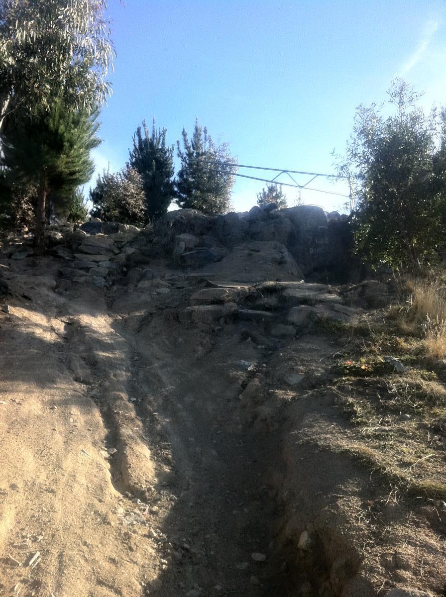 A dirt path leading up a rocky incline, surrounded by trees and clear blue skies. The trail shows signs of erosion and is lined with rocks, suggesting it is a natural or hiking route. Stromlo Forest Park mountain bike trail.