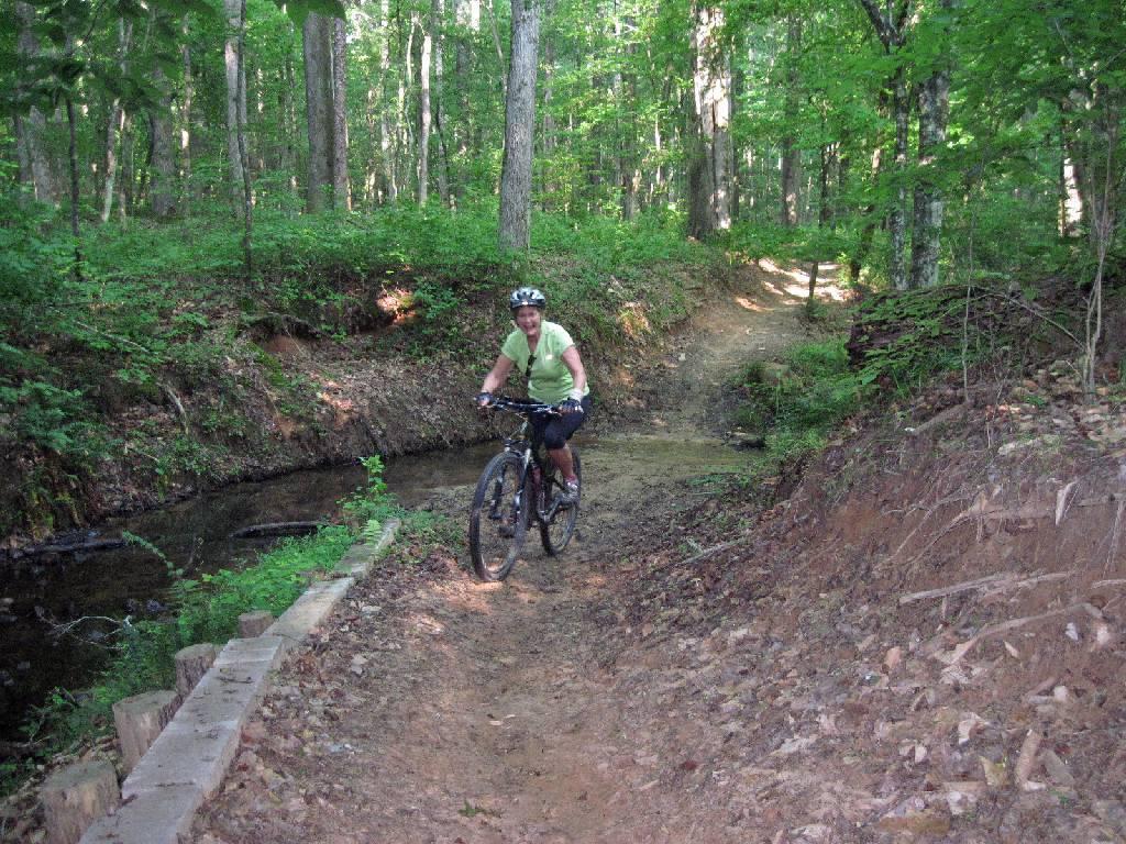 A person riding a mountain bike on a forest trail, with lush green trees and undergrowth surrounding them. The path includes a small wooden bridge over a shallow stream, and the rider is smiling while navigating the trail. Natural sunlight filters through the foliage, creating a vibrant outdoor setting. Long Cane Horse Trail mountain bike trail.