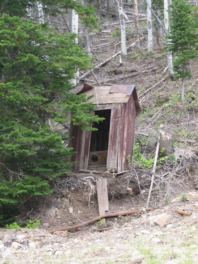 An old, weathered wooden outhouse stands on a hillside, partially surrounded by green foliage and trees. The structure has a rusty metal roof and an open door, revealing a simple interior. The ground around the outhouse is uneven, with exposed dirt and fallen branches, suggesting a secluded, rural location. Sweeney Switchbacks mountain bike trail.