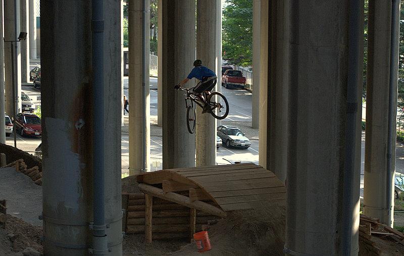 A cyclist in a blue shirt performs a jump off a wooden ramp located beneath a concrete structure, surrounded by tall columns. The scene captures the dynamic motion of the bike in mid-air against an urban backdrop with cars and greenery visible in the distance. I-5 Colonnade Mountain Bike Park mountain bike trail.
