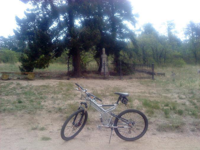 A silver mountain bike resting on a dirt path surrounded by grass and trees. In the background, a fenced area with a stone structure can be seen, partially obscured by trees. The sky is overcast, creating a moody atmosphere. Kipp