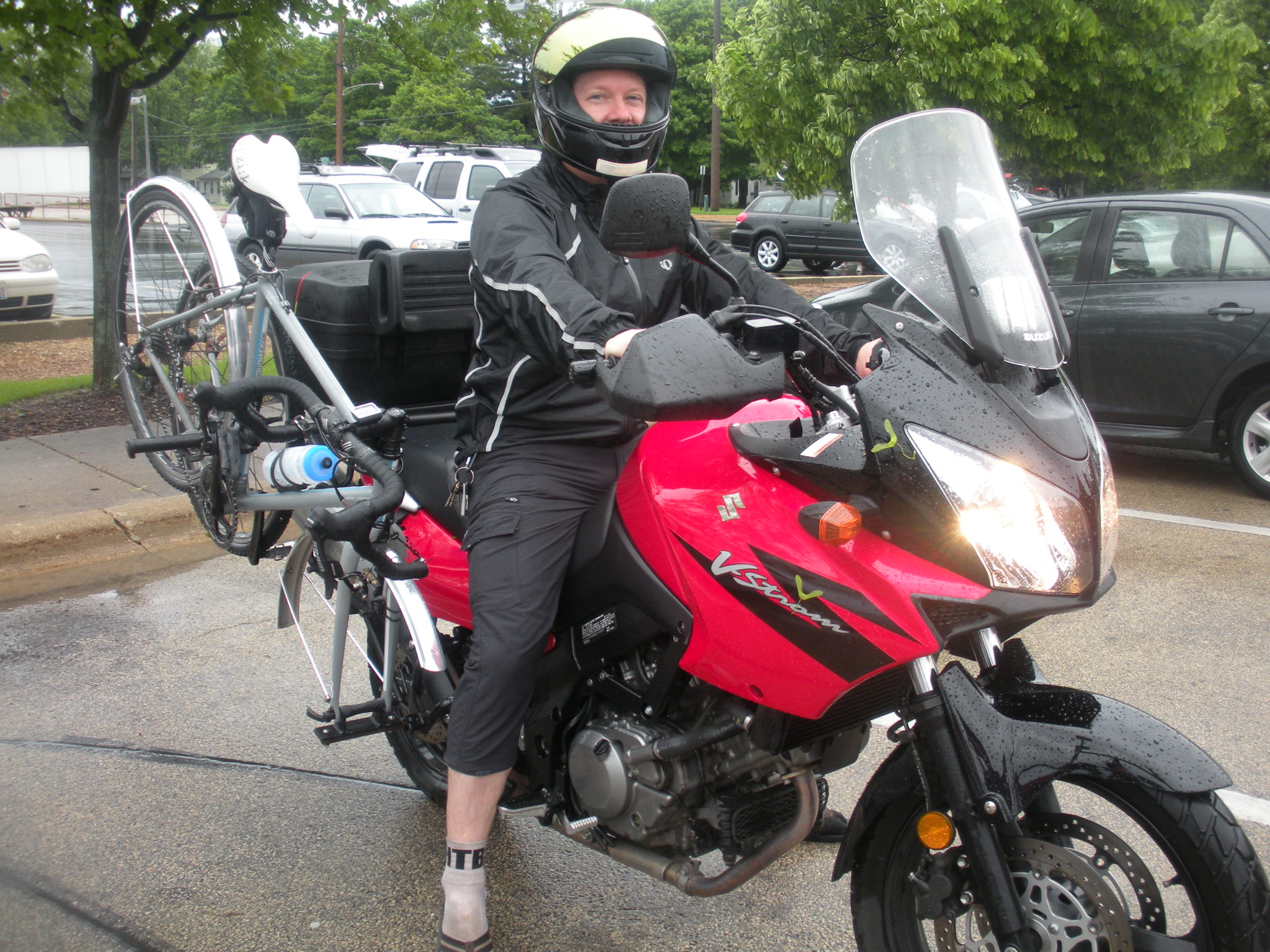 A person wearing a helmet and motorcycle gear sits on a red motorcycle, which has a bicycle attached to the back. The scene appears to be rainy, with wet pavement and greenery in the background. The motorcycle is parked in a lot with cars visible in the distance.