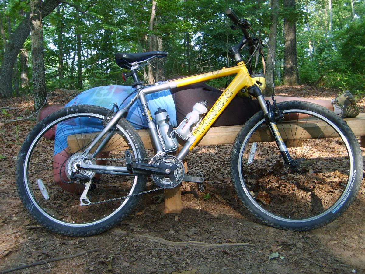 A yellow Trek mountain bike parked beside a wooden bench in a wooded area. The bike has a water bottle mounted and is positioned on a bed of leaves. In the background, there are trees and another person casually resting on the bench. Blankets Creek mountain bike trail.