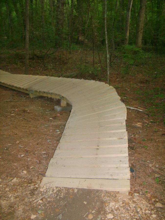 A wooden, curved pathway constructed in a forested area, surrounded by trees and underbrush. The path consists of slatted boards and is partially elevated above the ground, winding through the natural landscape. Blankets Creek mountain bike trail.