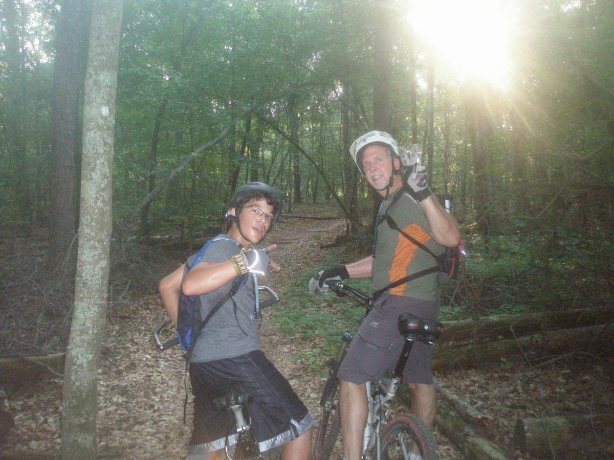 A boy and an adult man are posing for the camera while biking on a forest trail. Both are wearing helmets and biking gear. The boy gives a thumbs-up while slightly turning back to face the camera. The man is smiling and making a peace sign with his hand. Sunlight filters through the trees, creating a warm glow in the background. The trail is surrounded by lush green foliage and fallen leaves. Gainesville College mountain bike trail.