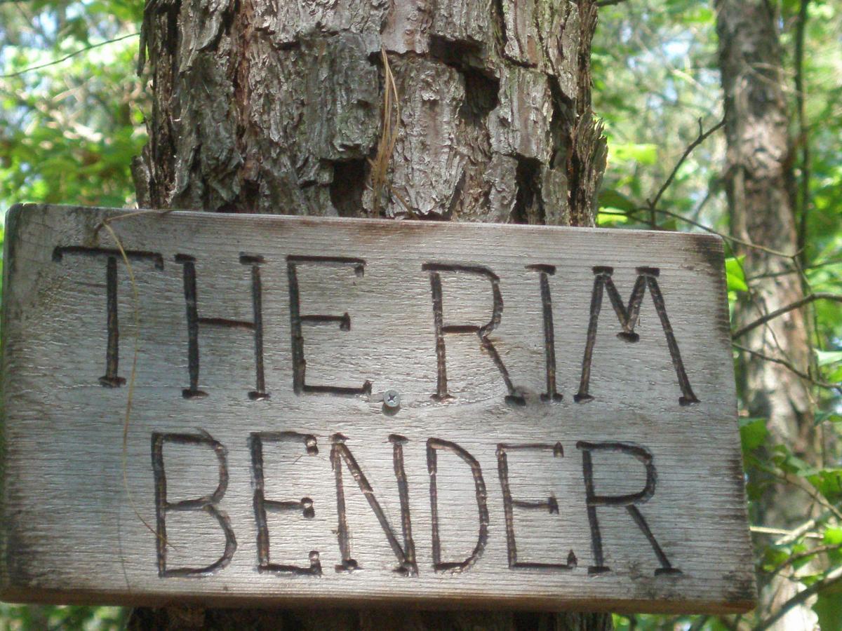 Wooden sign reading "THE RIM BENDER" mounted on a tree in a forested area, with green foliage in the background. Harbins Park mountain bike trail.