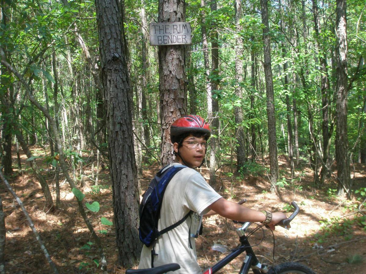 A young person wearing a red helmet and a light shirt is standing next to a mountain bike in a lush forest. In the background, there is a wooden sign labeled "The Rim Bender" attached to a tree. The area is surrounded by tall trees and underbrush, indicating a mountain biking trail. Harbins Park mountain bike trail.