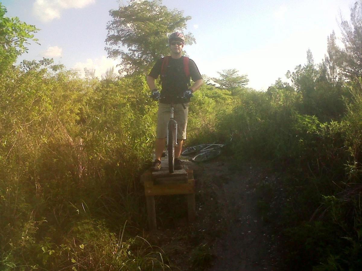 A person standing on a wooden platform with one foot on a mountain bike tire, surrounded by greenery and a dirt trail. The individual is wearing a helmet, gloves, and casual biking attire, with a bicycle resting nearby. The scene is set in a natural outdoor environment during daylight. West Delray Regional Park mountain bike trail.