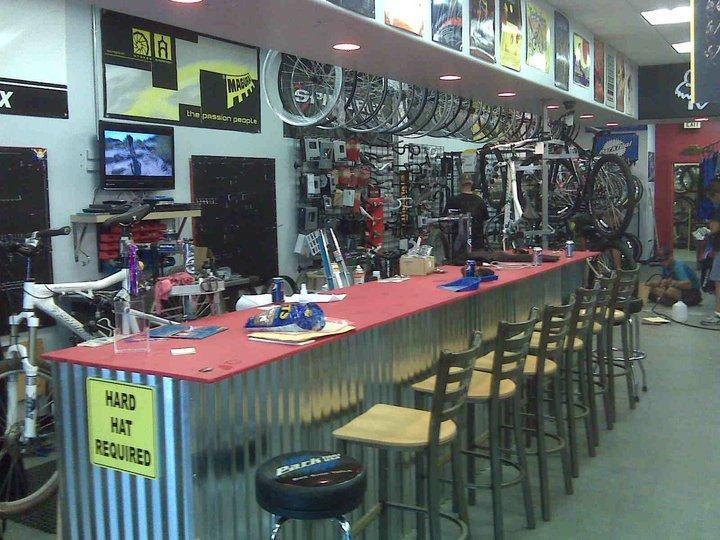 An interior view of a bicycle shop featuring a long red work counter with several stools, surrounded by shelves stocked with bike parts and accessories. The walls are adorned with various bike frames and artwork, and a TV is mounted on the wall displaying bike-related footage.